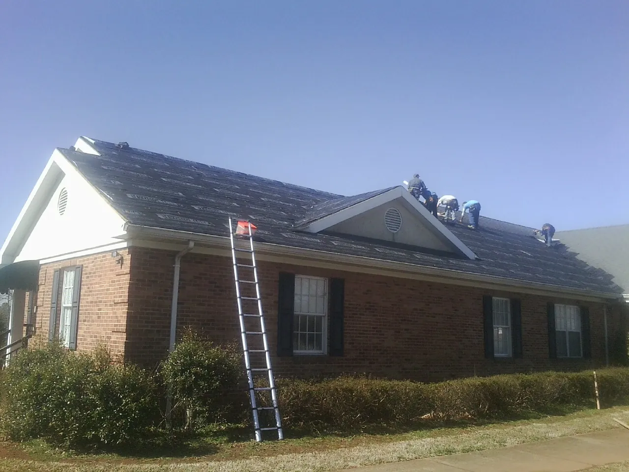Crew tearing shingles off a brick ranch roof with a ladder set — Emergency Roof Repair work by our crew in Ragland