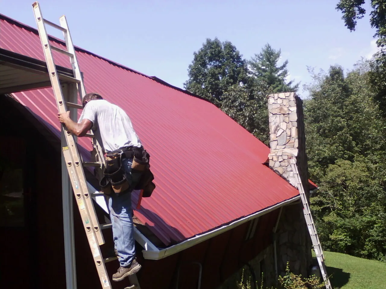 Roofer on a ladder beside a red metal roof and stone chimney — Soffit & Fascia Repair by Roofing Brothers in Ragland